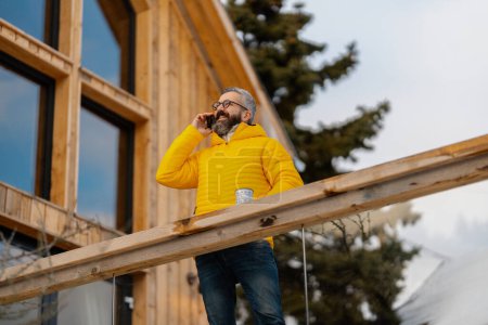 Photo for Mature man standing on cabins patio, drinking hot tea, coffee and enjoying beautiful winter day. Handsome man spending relaxing, stress-free winter weekend in cabin in mountains, enjoying alone time. - Royalty Free Image