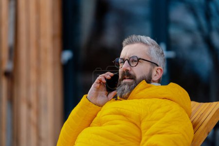 Photo for Portrait of handsome man phone calling from patio, during cold winter day. Handsome man spending relaxing, stress-free winter weekend in cabin in mountains, enjoying alone time. - Royalty Free Image