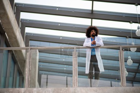 Téléchargez les photos : Portrait du médecin masculin en manteau blanc debout à l'hôpital, appuyé sur une balustrade en verre, regardant la caméra. - en image libre de droit