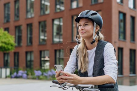 Téléchargez les photos : Belle femme d'âge moyen se déplacer à travers la ville en vélo, tenant smartphone. Femme banlieue de la ville voyageant du travail à vélo après une longue journée de travail. - en image libre de droit