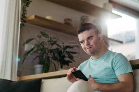 Téléchargez les photos : Portrait de jeune homme trisomique assis dans la cuisine avec smartphone à la main. - en image libre de droit
