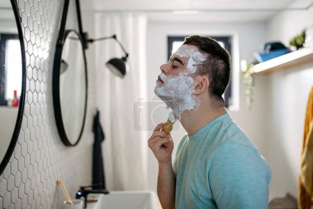 Photo for Young man with down syndrome learning how to shave, applying shaving foam, cream all over his face, looking at mirror. - Royalty Free Image