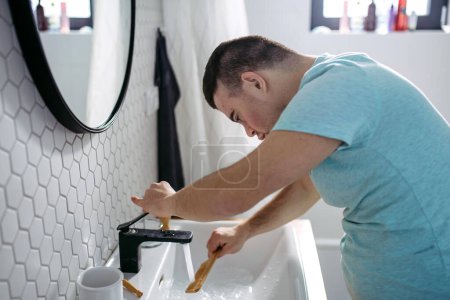 Téléchargez les photos : Vue latérale du jeune homme avec syndrome du duvet dans la salle de bain, peigne de nettoyage, rinçage dans l'eau. - en image libre de droit