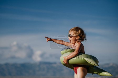 Téléchargez les photos : Vue latérale de la petite fille surprise à la plage avec anneau de jouet gonflable, pointant du doigt à distance. - en image libre de droit