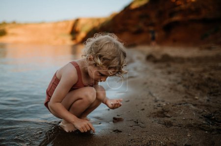 Foto de Niña en traje de baño jugando en la playa, agachándose y seraching para conchas en la arena. - Imagen libre de derechos