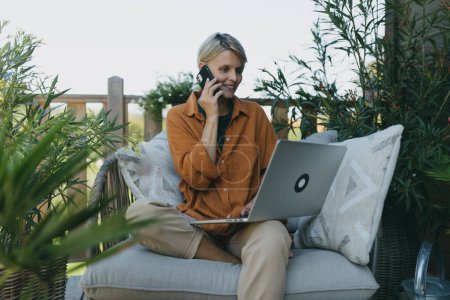 Photo for Woman working in garden, with laptop on legs. Businesswoman working remotely from outdoor homeoffice, making call on smartphone. Concept of outdoor home office. - Royalty Free Image