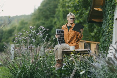 Photo for Woman in garden holding solar panel and tablet. Looking for solar panels grants, funds for homeowners. Solar energy and sustainable lifestyle for the family in house. - Royalty Free Image