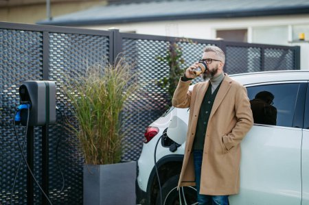 Retrato del hombre de negocios esperando mientras el coche eléctrico se está cargando, apoyándose en el vehículo, desplazándose en su teléfono inteligente y bebiendo café. Una estación de carga de vehículos eléctricos en casa.
