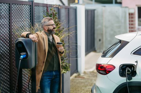 Portrait of businessman waiting while electric car is charging, leaning against the vehicle, scrolling on his smartphone, and drinking coffee. An electric vehicle charging station at home.