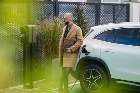 Photo for Businessman charging electric car before going to office. An electric vehicle charging station in front of the office building. Charging at work, workplace. - Royalty Free Image