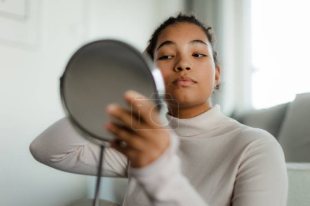Foto de Retrato de hermosa mujer con curvas, mirando pequeño espejo de maquillaje redondo en casa. Régimen de cuidado de la piel. - Imagen libre de derechos