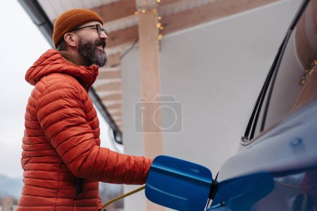 Photo for Man charging electric car during cold snowy day. Hansome mature man putting charger in charging port during cold weather. Charging and driving electric vehicles during winter season. Low angle shot. - Royalty Free Image