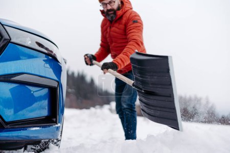 Téléchargez les photos : Un homme libérant sa voiture coincée dans la neige avec une pelle, sortant la voiture de la dérive de neige. Ramasser et jeter de la neige, pelleter. - en image libre de droit