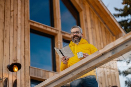 Photo for Mature man standing on cabins patio, drinking hot tea, coffee and reading book. Handsome man spending relaxing, stress-free winter weekend in cabin in mountains, enjoying alone time. - Royalty Free Image