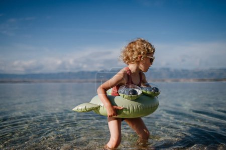 Téléchargez les photos : Vue du dessus de la petite fille en maillot de bain rose à la plage avec anneau de jouet gonflable, jouant dans l'eau regardant la mer. - en image libre de droit