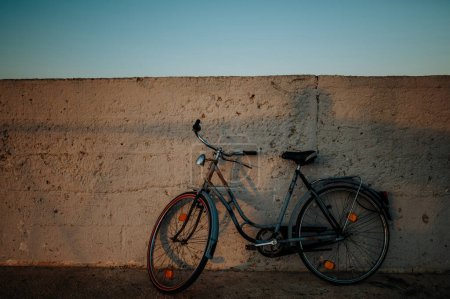 Téléchargez les photos : Vieux vélo appuyé contre un mur de béton, rayons du soleil couchant et ombres. Scène prise avec espace de copie. - en image libre de droit