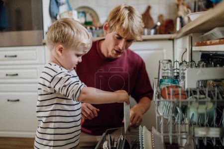 Photo for Little boy helping father to load dishwasher after family breakfast. Cleaning kitchen before leaving to work and daycare. Family morning routine. - Royalty Free Image