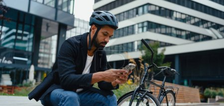 Téléchargez les photos : Commutateur de la ville, assis sur le banc, défilant sur le smartphone. Homme d'affaires voyageant du travail à la maison à vélo. Style de vie urbain, bannière avec copyspace. - en image libre de droit