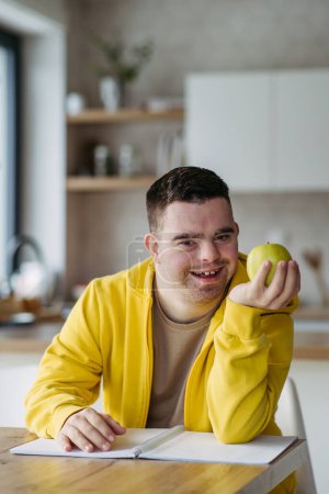 Téléchargez les photos : Portrait d'un jeune homme trisomique tenant une pomme, assis à la table. - en image libre de droit