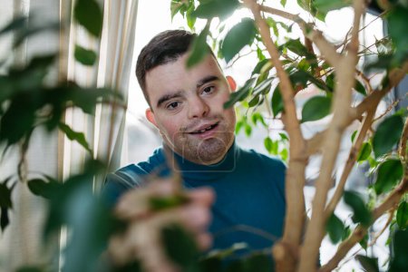 Photo for Portrait of young man with Down syndrome in the middle of houseplants. Taking care of indoor plant, touching and snuggling plant leaf. - Royalty Free Image