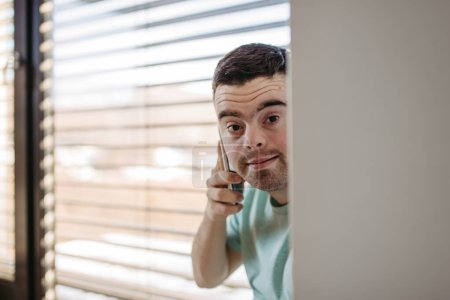 Téléchargez les photos : Portrait de jeune homme trisomique assis par la fenêtre, faire un appel téléphonique avec smartphone. - en image libre de droit