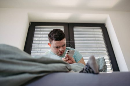Téléchargez les photos : Jeune homme avec le syndrome du duvet couché dans son lit, regardant smartphone le matin. routine du matin pour les hommes handicapés. - en image libre de droit