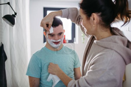 Photo for Mother applying shaving foam on sons face. Young man with down syndrome learning how to shave. Concept of mothers day and motherly love. - Royalty Free Image