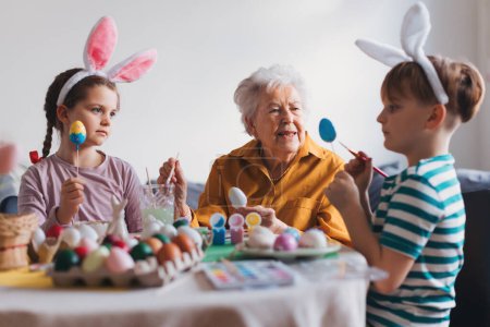 Foto de Abuela con niños pequeños decorando huevos de Pascua en casa. Tradición de pintar huevos con pincel y tinte de huevo de Pascua. Concepto de vacaciones familiares de Pascua. - Imagen libre de derechos