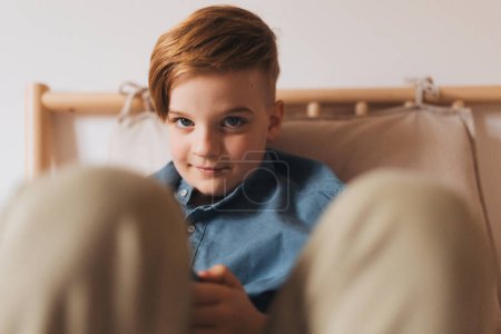 Portrait of young boy sitting on bed, looking at camera with soft smile. Handsome young boy in bedroom.