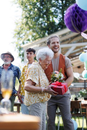 Photo for Garden birthday party for senior lady. Beautiful senior birthday woman receiving birthday wishes and gifts from grandson. - Royalty Free Image