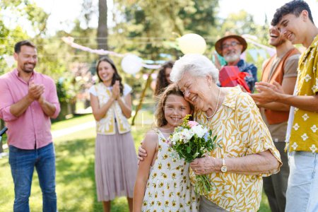 Foto de Fiesta de cumpleaños en el jardín para la señora mayor. Hermosa mujer de cumpleaños mayor que recibe flores de su nieta. - Imagen libre de derechos