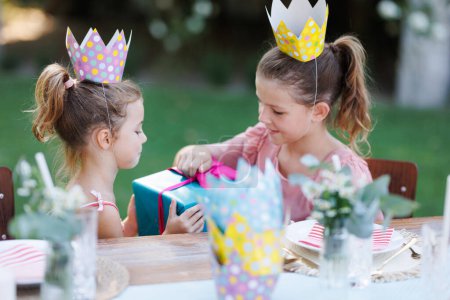 Photo for Little friends at a birthday party outdoors at the garden. Cute girls with paper crowns opening birthday gift. Birthday garden party for children. - Royalty Free Image