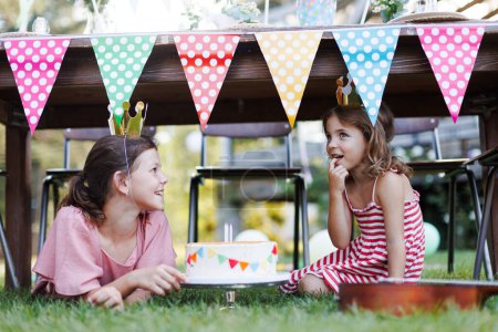 Téléchargez les photos : Filles mignonnes avec des couronnes en papier manger gâteau d'anniversaire sous la table. Fête d'anniversaire pour les enfants. Fête d'anniversaire jardin pour les enfants. - en image libre de droit