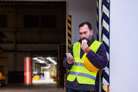 Photo for Portrait of worker taking break from work, eating sandwich, scrolling standing on loading dock, outdoors. Eating light lunch outdoors. - Royalty Free Image