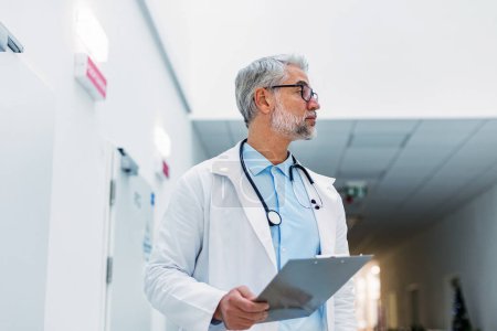 Portrait of mature doctor in hospital corridor. Handsome doctor with gray hair wearing white coat, stethoscope around neck standing in modern private clinic.