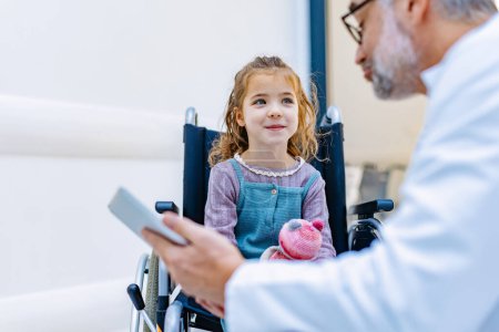 Photo for Friendly pediatrician talking to little patient in wheelchair. Cute preschool girl in wheelchair greeting doctor in hospital. Concept of children healthcare and emotional support for child patients. - Royalty Free Image