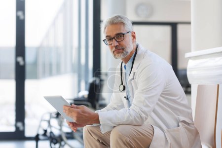 Portrait of smiling doctor sitting in hospital corridor. Handsome doctor with gray hair wearing white coat, stethoscope around neck standing in modern private clinic and looking at camera.