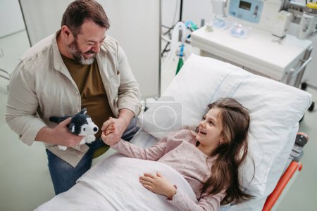 Foto de Padre sentado junto a su hija acostada en la cama del hospital, jugando con un juguete de peluche. Regalo de papá a una niña enferma en el hospital. - Imagen libre de derechos