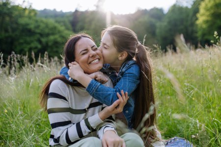 Beautiful mother with daughter, embracing or hugging, sitting in the grass at meadow. Concept of Mothers Day and maternal love.