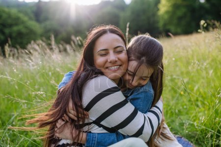 Beautiful mother with daughter, embracing or hugging, sitting in the grass at meadow. Concept of Mothers Day and maternal love.