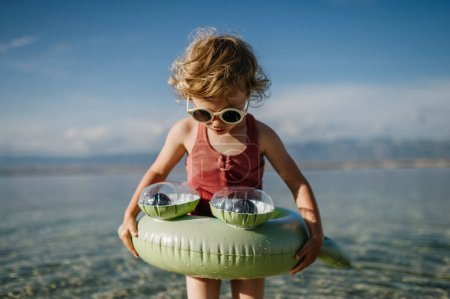 Foto de Vista superior de niña pequeña en traje de baño rosa en la playa con anillo de juguete inflable, jugando en el agua mirando al mar. - Imagen libre de derechos