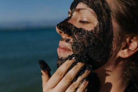 Woman applies healing mud to her face and body. Natural healing mud in Croatia on the beach. Therapeutic mud or peloids rich in minerals and organic substances.