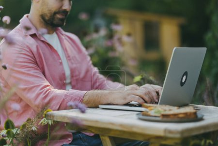 Photo for Man working outdoors in the garden, eating lunch or snack, sandwich. Businessman working remotely from homeoffice, thinking about new business or creative idea. - Royalty Free Image