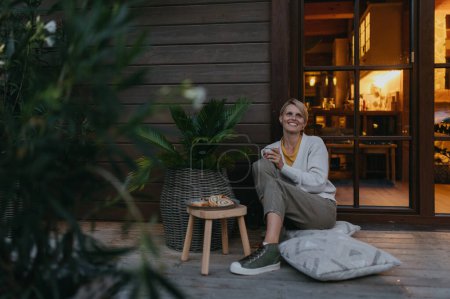 Photo for Woman sitting on the terrace during dusk, sipping warm tea and enjoying the view. Inside the house, its warm and inviting, while she is dressed in a cozy sweater, sitting on soft pillows. Hygge - Royalty Free Image