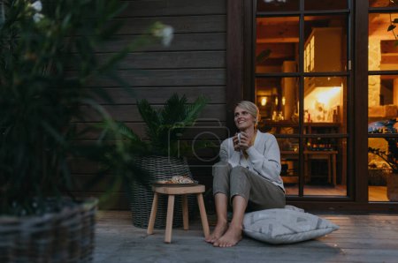 Téléchargez les photos : Femme assise sur la terrasse au crépuscule, sirotant du thé chaud et profitant de la vue. A l'intérieur de la maison, son chaleureux et accueillant, alors qu'elle est habillée d'un pull confortable, assis sur des oreillers doux. Hygge - en image libre de droit