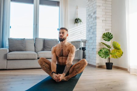 Foto de Hombre guapo haciendo yoga, estirándose, descansando después del entrenamiento en casa, resoluciones de Año Nuevo, ejercitándose como estilo de vida saludable y autocuidado. Concepto de rutina de entrenamiento matutino o nocturno. - Imagen libre de derechos