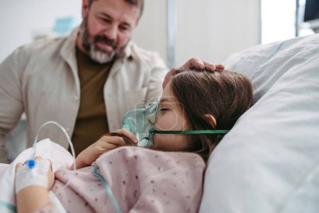 Téléchargez les photos : Père tenant la main de sa fille dans son lit d'hôpital. Enfant patient dans un lit d'hôpital avec un masque à oxygène sur le visage en soins intensifs. - en image libre de droit