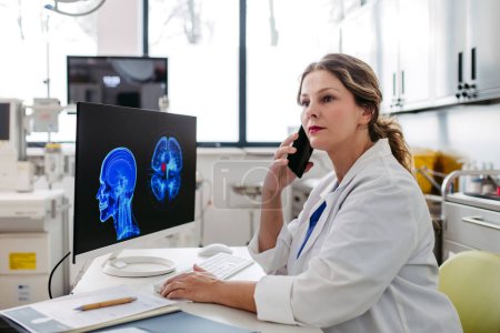 Photo for Female doctor working on computer in doctors office, looking at MRI scan, phone calling test results to patient. Doctor consulting scan with the other doctors. Telephone consultations. - Royalty Free Image