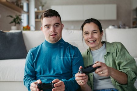 Foto de Retrato de un joven con síndrome de Down con su madre jugando videojuegos, sosteniendo controladores de juego. Concepto de amor y crianza de los hijos discapacitados. - Imagen libre de derechos
