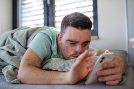 Foto de Hombre joven con síndrome de Down acostado en su cama, mirando el teléfono inteligente por la mañana. Rutina matutina para hombre con discapacidad. - Imagen libre de derechos